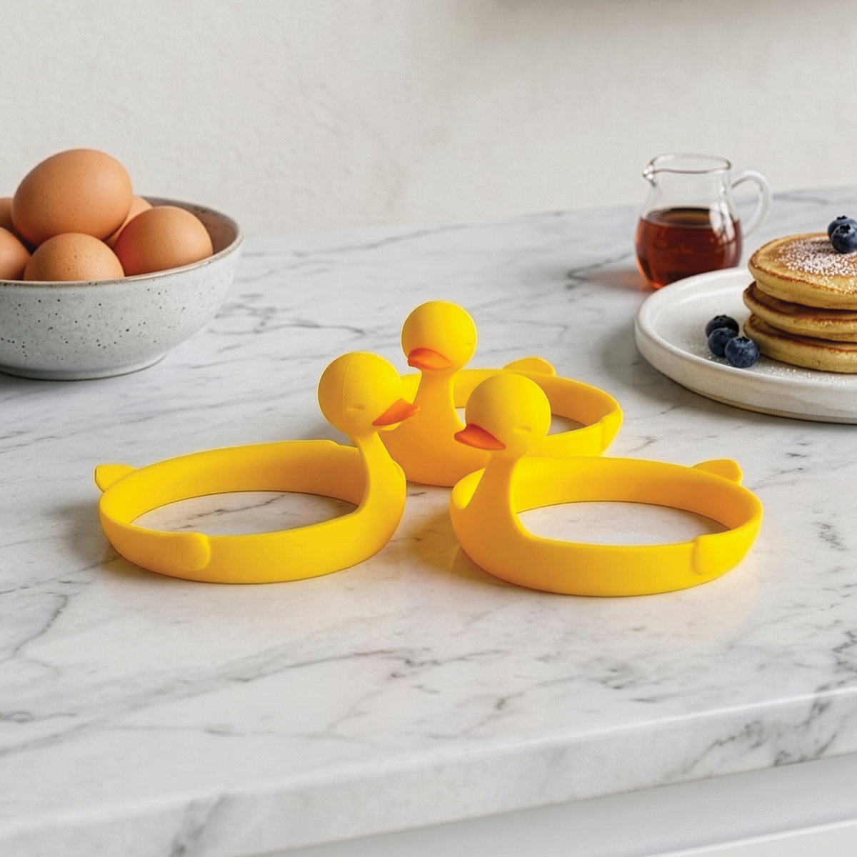 Three Sunny Side Duck egg rings sit on marble near eggs, pancakes, and syrup.